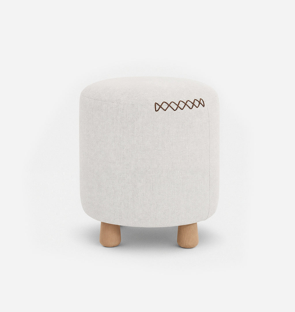 A round upholstered ottoman stool with a white textured linen fabric with leather x-stitching and three short natural wood legs, shown against a plain white background.