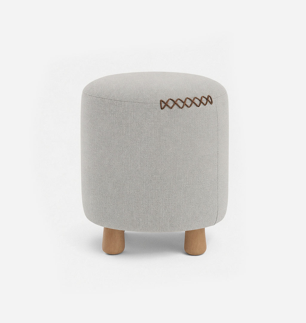 A round upholstered ottoman stool with a light beige textured linen fabric with leather x-stitching and three short natural wood legs, shown against a plain white background.