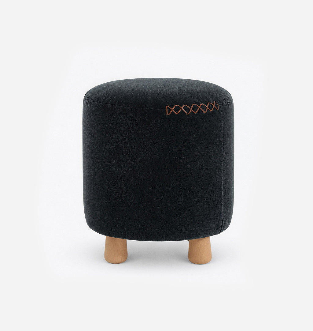 A round upholstered ottoman stool with a navy velvet fabric with leather x-stitching and three short natural wood legs, shown against a plain white background.