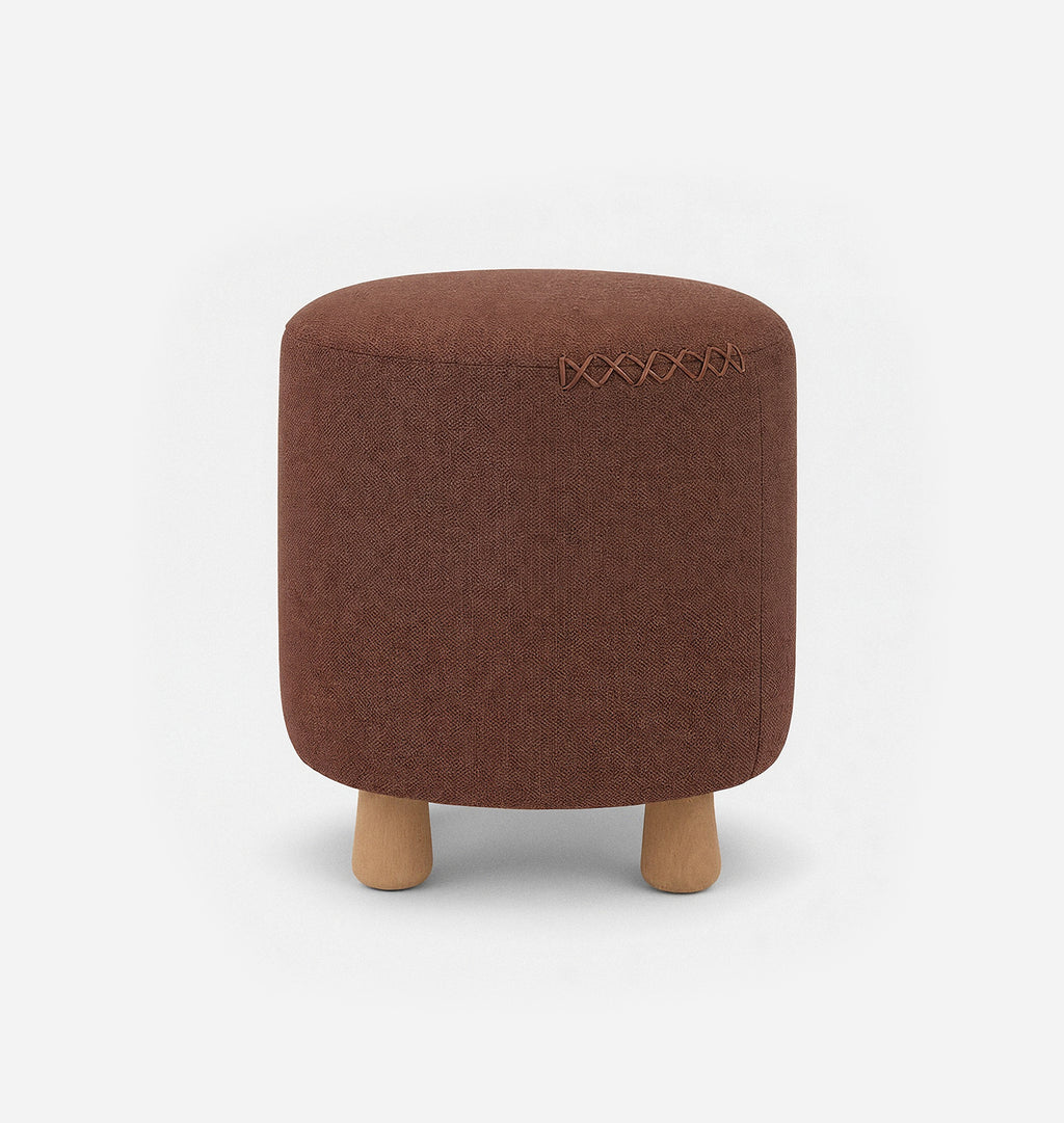 A round upholstered ottoman stool with a reddish brown textured linen fabric with leather x-stitching and three short natural wood legs, shown against a plain white background.