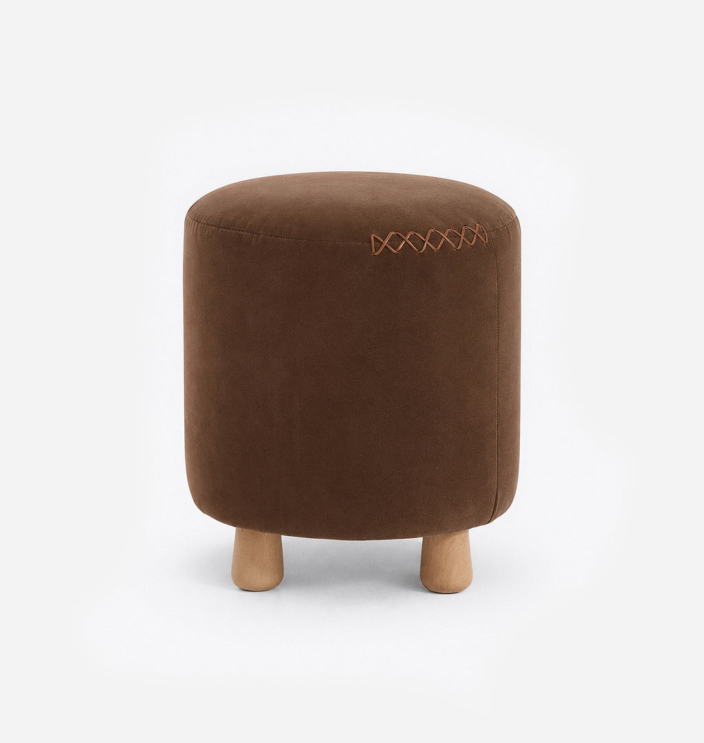 A round upholstered ottoman stool with a brown velvet fabric with leather x-stitching and three short natural wood legs, shown against a plain white background.