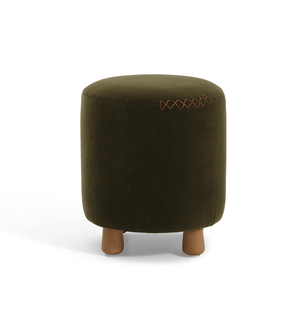 An olive green velvet cylindrical ottoman with short wooden legs and a decorative leather x-stitching on the upper right side, shown against a plain white background.