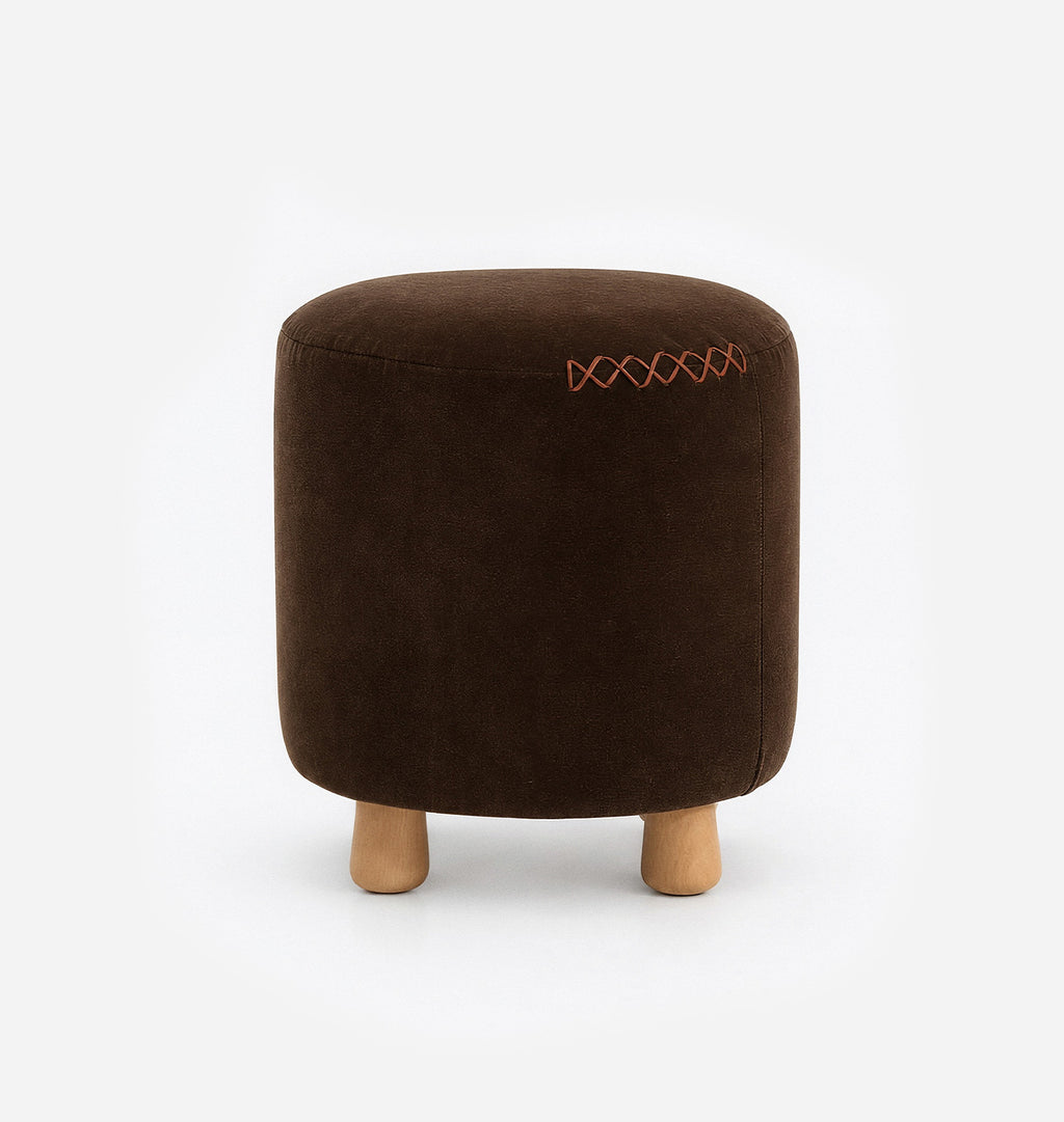 A round upholstered ottoman stool with a dark brown velvet fabric with leather x-stitching and three short natural wood legs, shown against a plain white background.