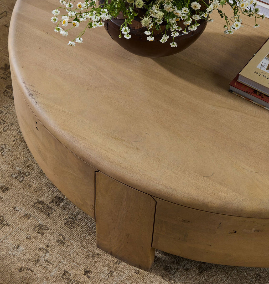 A close-up of a round, light oak wood coffee table with a unique angled leg design, styled with a dark brown vase of white daisies and books on a patterned beige rug.
