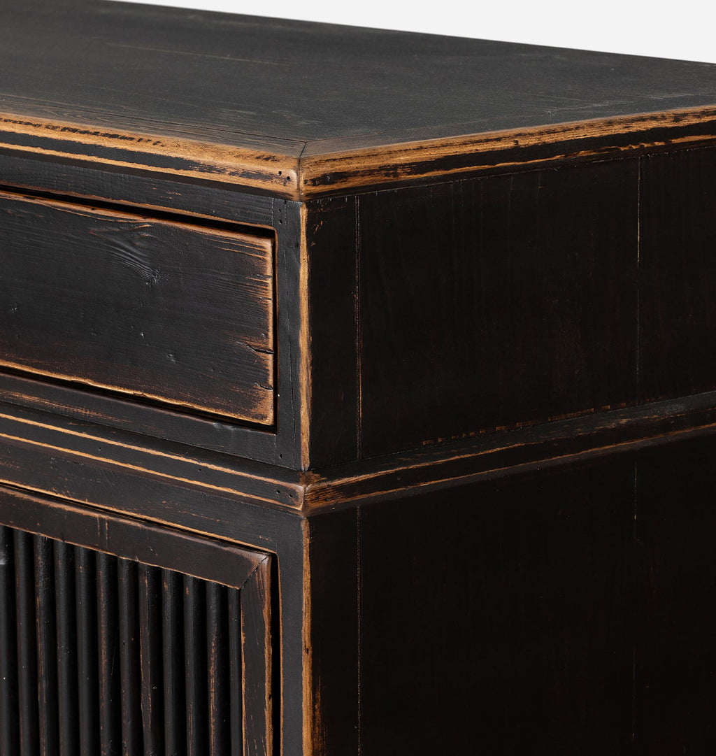 Close-up of a dark distressed wooden cabinet corner, showing a black finish with warm brown wood showing through the worn edges and a slatted door panel on the lower left.