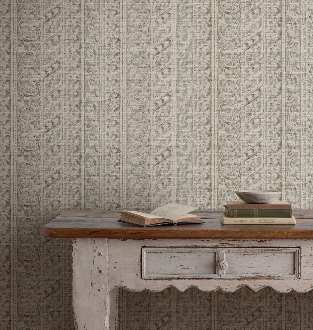 Java Grasscloth Wallpaper with a vertical ornate pattern in muted sage green, beige, and gray tones, styled behind a rustic white-painted wooden table with books and a bowl.