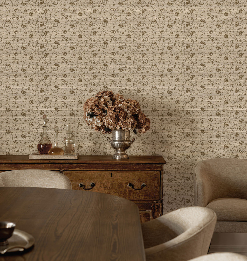 A styled interior scene showing Margueritte Grasscloth Wallpaper in a warm beige and brown pattern, a dark wood sideboard with a silver vase of dried flowers, dining chairs, and part of a table in a real room setting.