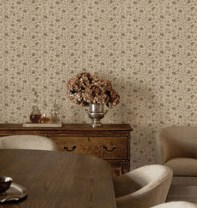 A styled interior scene showing Margueritte Grasscloth Wallpaper in a warm beige and brown pattern, a dark wood sideboard with a silver vase of dried flowers, dining chairs, and part of a table in a real room setting.