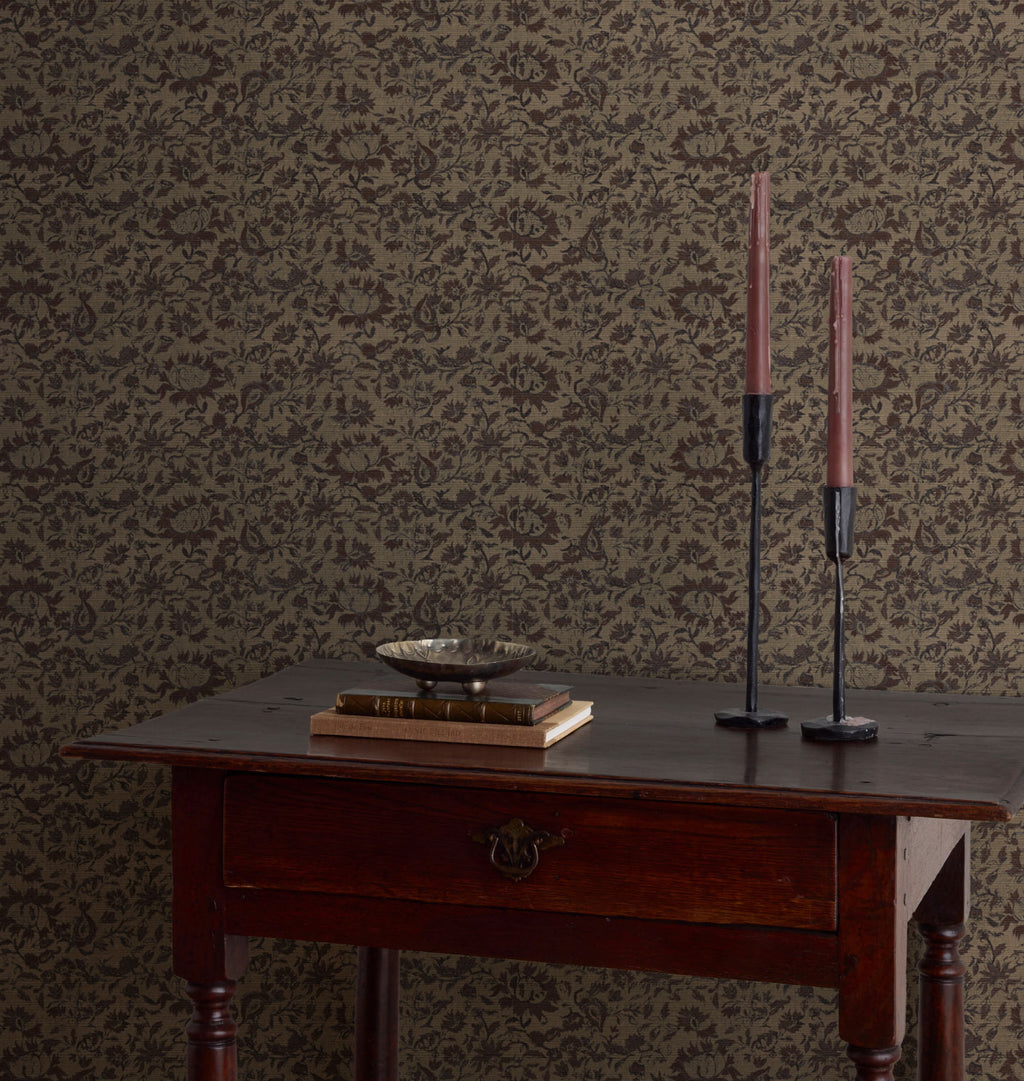 Damask patterned Raffi Grasscloth Wallpaper in dark brown and beige, styled behind a dark wood table with books, a bowl, and two tall pink candles.