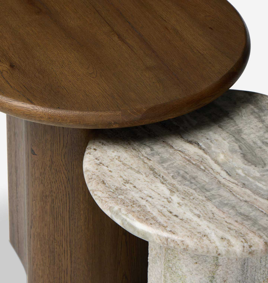 Close-up of a round wooden side table partially overlapping a round marble side table with a beige and gray veined stone top.
