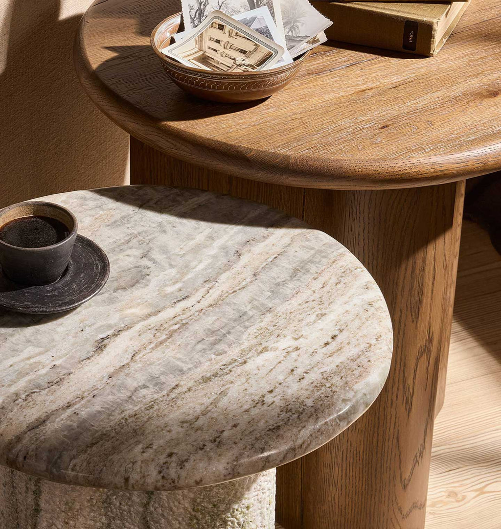 A close-up of a round side table with a polished light beige marble top, shown in a warm, sunlit room setting alongside a wooden table.