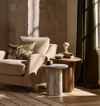 A beige upholstered armchair in a sunlit room with a patterned green and beige throw pillow, a round wood end table with books and a small bust, and a stone-textured side table holding a cup, set against a stone wall and dark curtain.
