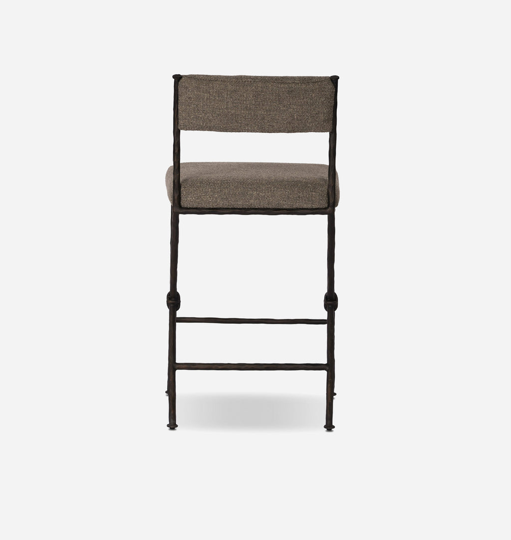 A modern bar stool with a textured taupe fabric seat and backrest, supported by a dark metal frame with decorative turned leg caps and a lower support rail, shown against a plain white background.