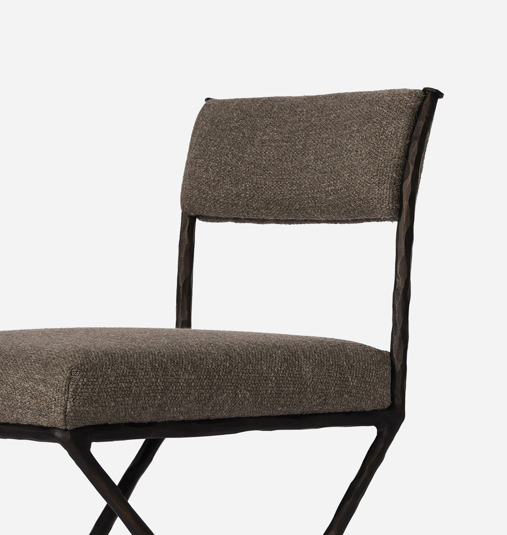 A close-up of a modern accent chair featuring textured taupe-brown woven upholstery on the seat and backrest, supported by a dark metal frame with a twisted side rail and crisscrossing legs.