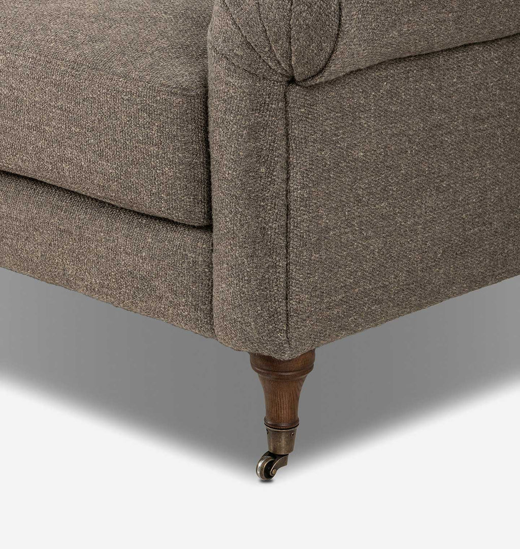 Close-up of the corner of a upholstered sofa in textured taupe fabric, showing the rolled arm, cushion seam, turned wooden leg, and small metal caster wheel.