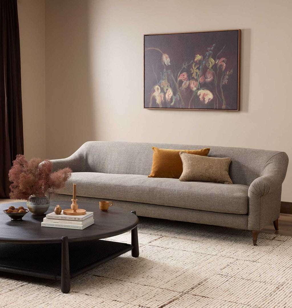 A gray upholstered sofa in a styled living room, with two accent pillows, a dark round coffee table, and a framed floral painting on the beige wall behind it.