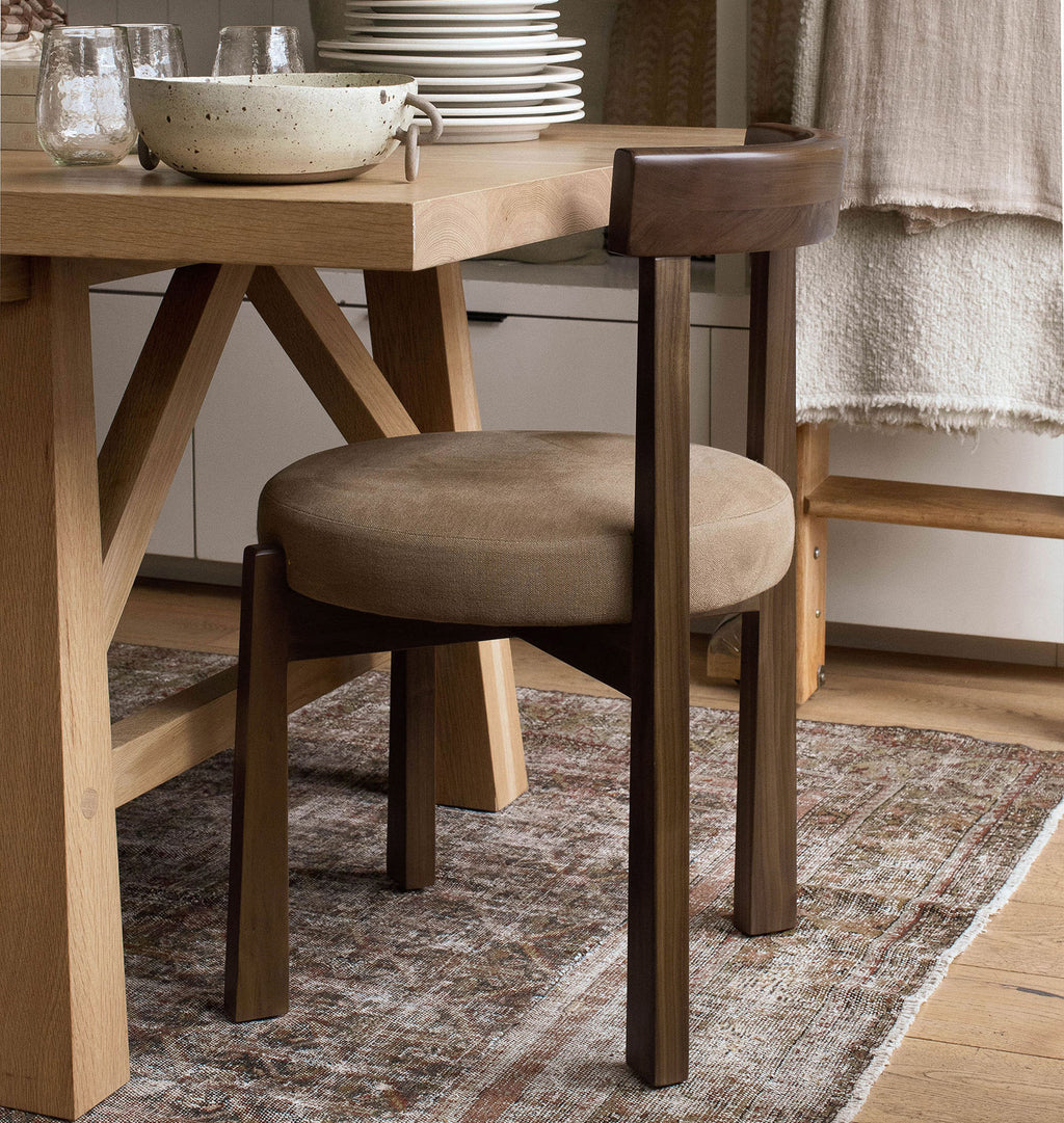 A round upholstered dining chair with a dark wood frame and seat, shown beside a wooden dining table in a real interior setting with a rug, bowls, plates, and a draped textile.
