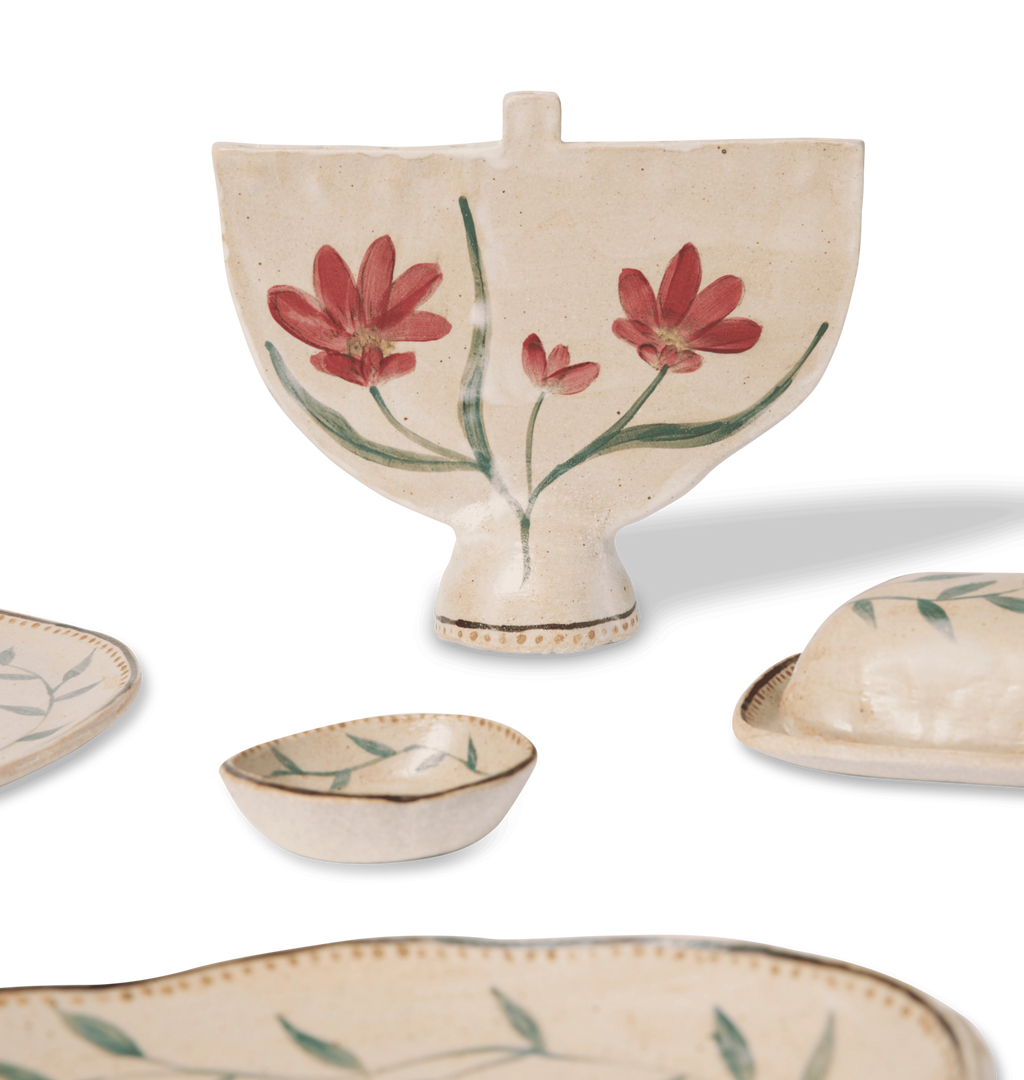 A collection of hand-painted ceramic dishes on a white background, featuring a central cream-colored menorah with red flowers and green leaves, surrounded by matching plates, a small bowl, and a lidded dish.