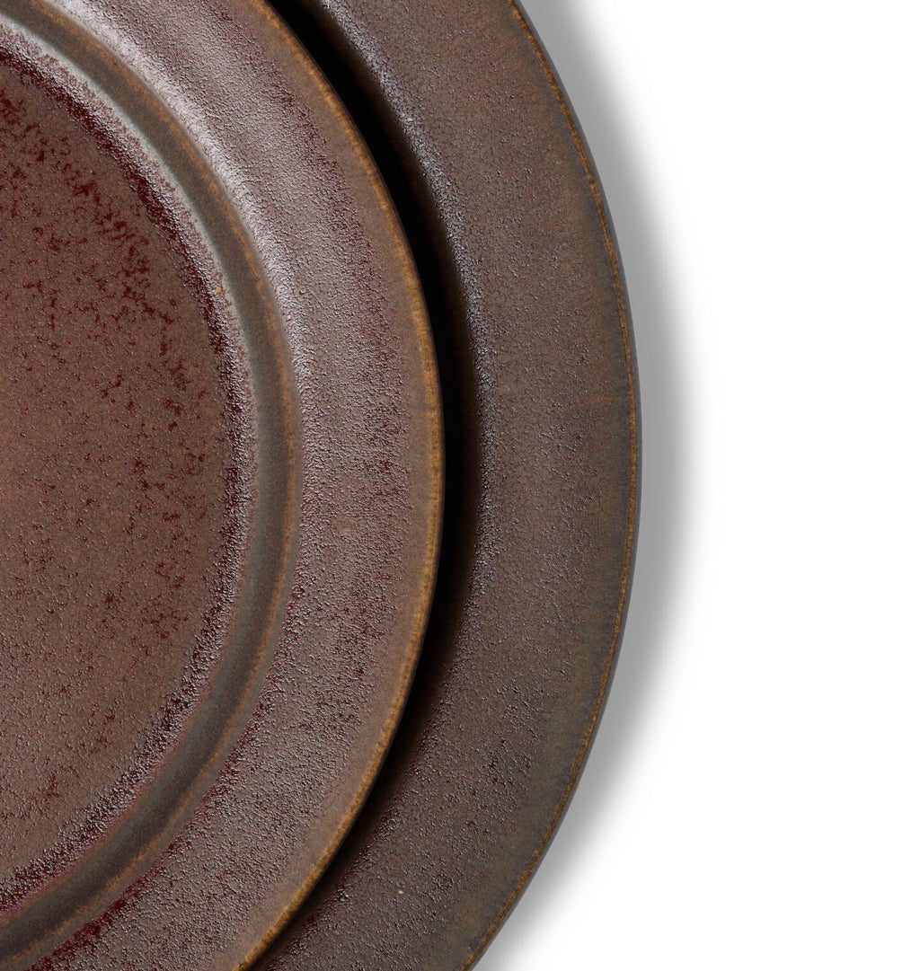 A close up detail shot of a round ceramic salad plate stacked on a dinner plate in a dark reddish brown glaze shown on a white background
